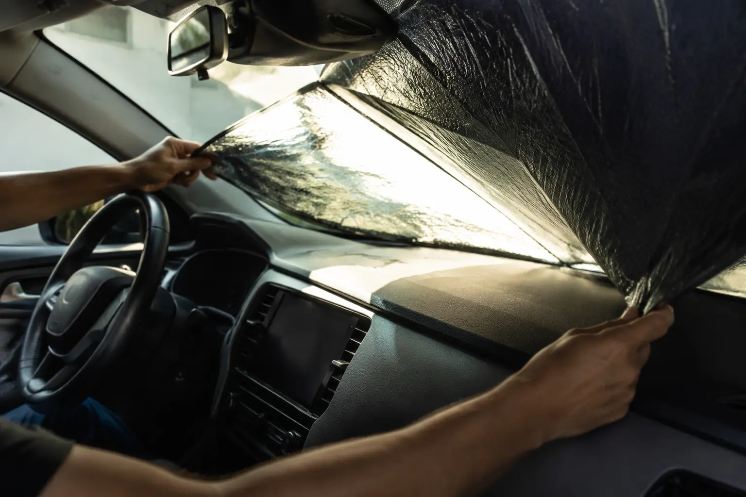 Adult hands unfolding a plain reflective windshield sun screen inside an unbranded car